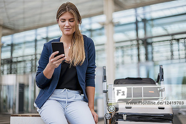 Smiling young businesswoman sitting outdoors with cell phone and suitcase