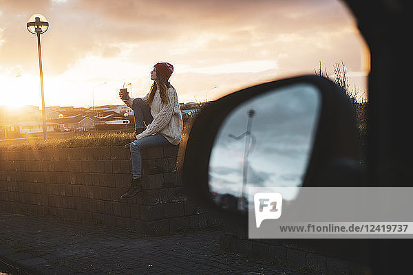 Iceland  young woman with coffee to go at sunset  wing mirror