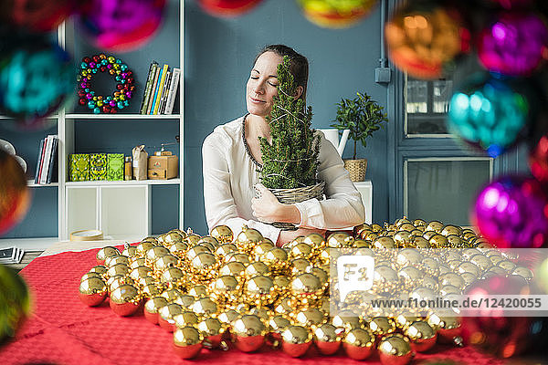 Woman sitting at table with many golden Christmas baubles holding potted fir tree
