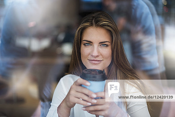 Portrait of smiling young woman behind windowpane with takeaway coffee