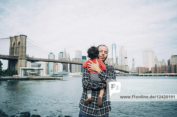 USA  New York  New York City  Father hugging a baby in Brooklyn with Brooklyn Bridge and Manhattan in the background