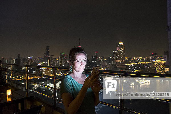 Thailand  Bangkok  portrait of woman standing on roof terrace by night using cell phone