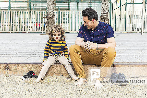 Spain  Barcelona  father and son sitting next to the beach barefoot