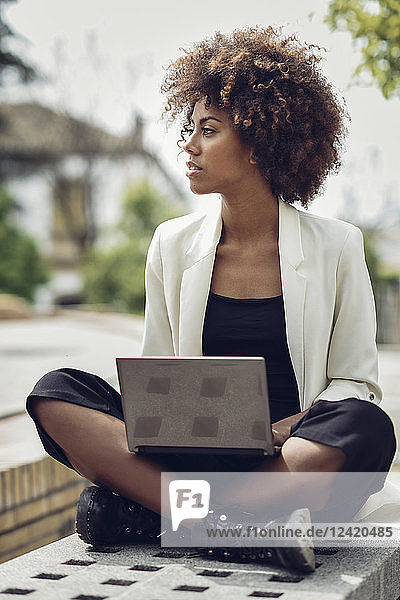 Fashionable young woman with curly hair sitting on bench with laptop