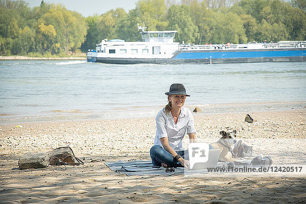Smiling woman sitting on blanket at a river with dog using laptop
