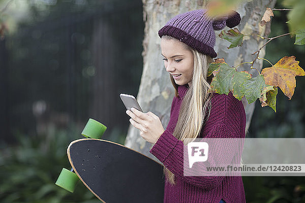 Smiling teenage girl looking at cell phone holding skateboard