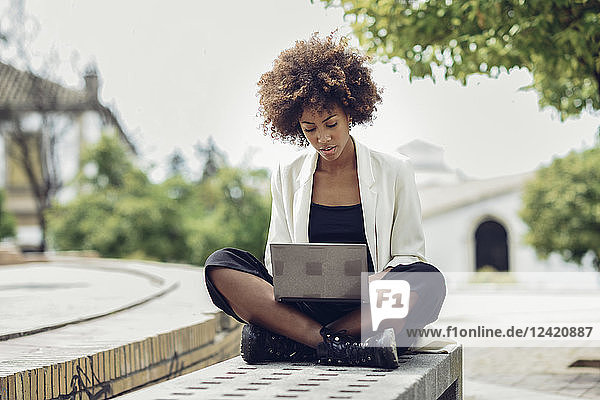 Fashionable young woman with curly hair sitting on bench using laptop