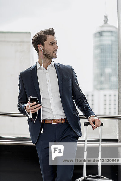 Young businessman at the window with cell phone  earbuds and rolling suitcase