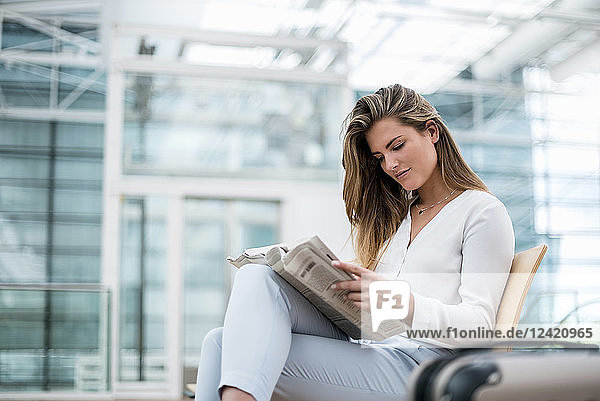 Young businesswoman sitting outdoors with suitcase reading newspaper