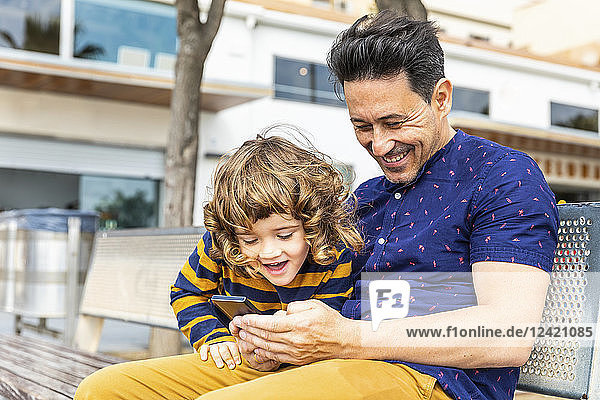 Spain  Barcelona  happy father and son with a smartphone sitting on bench