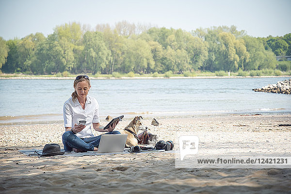 Woman sitting on blanket at a river with dog using portable devices