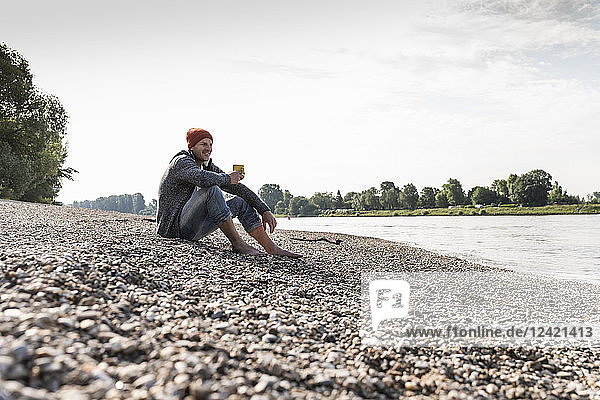 Mature man wearing red beanie and drinking coffee at Rhine riverbank