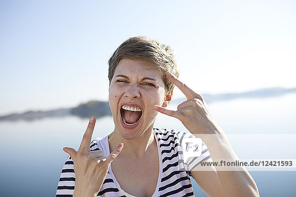 Portrait of screaming woman in front of lake showing Rock And Roll Sign