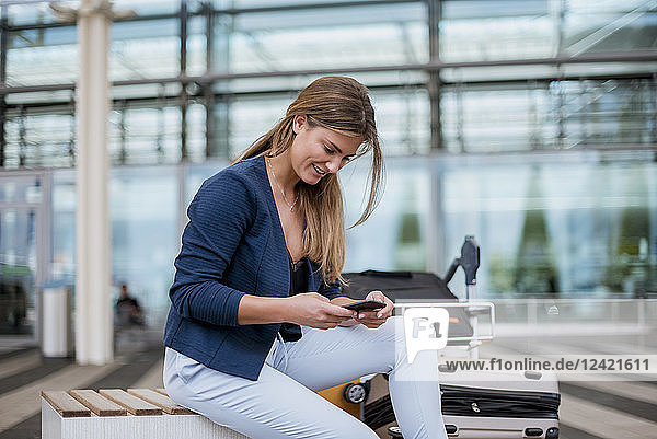 Smiling young businesswoman sitting outdoors with cell phone and suitcase
