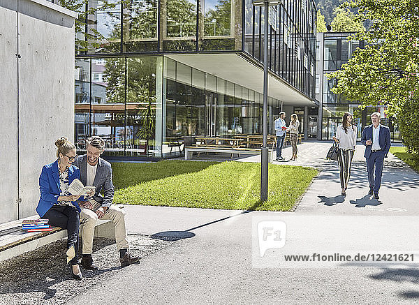 Smiling colleagues with book sitting on bench outside office building
