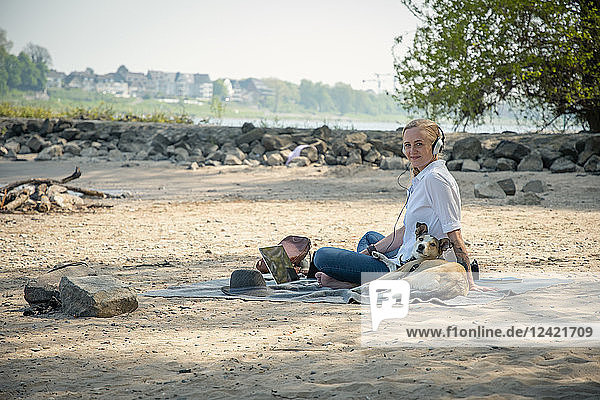 Smiling woman sitting on blanket at a river with dog wearing headphones