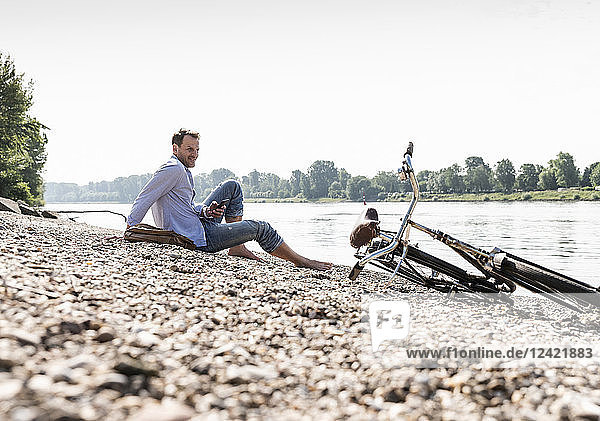 Mature man with bike and smartphone sitting at Rhine riverbank