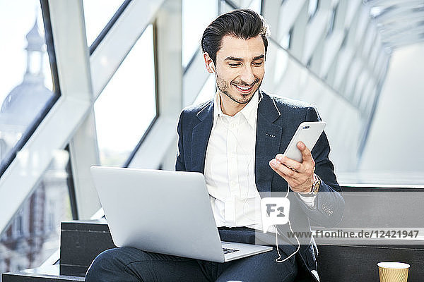 Smiling businessman sitting on stairs wearing earphones using cell phone and laptop