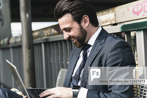 Happy businessman sitting outdoors on a bench using laptop