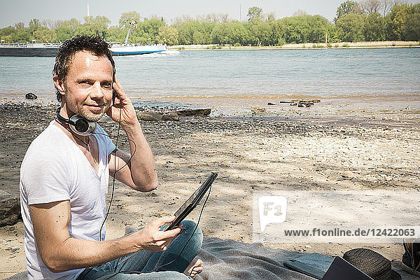 Portrait of smiling man sitting on blanket at a river using tablet