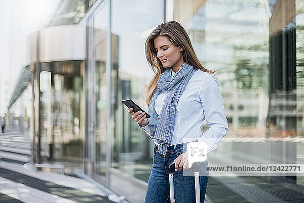Young businesswoman with suitcase looking at smartphone