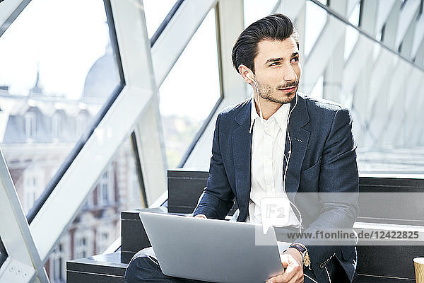 Businessman sitting on stairs wearing earphones and using laptop