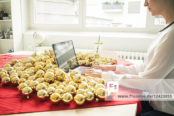 Woman sitting at table with many golden Christmas baubles working on laptop