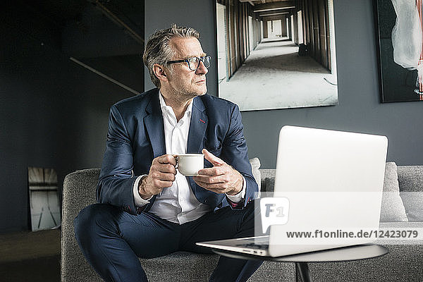 Mature businessman sitting on couch with cup of coffee and laptop