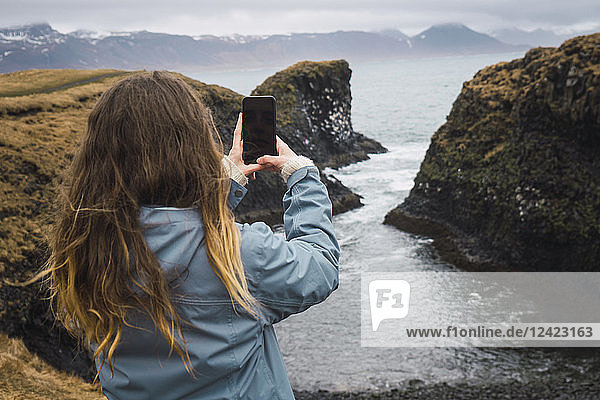 Iceland  back view of young woman taking picture with smartphone at coast