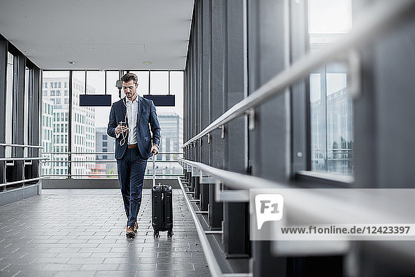 Young businessman in a passageway with cell phone  earbuds and rolling suitcase on the go