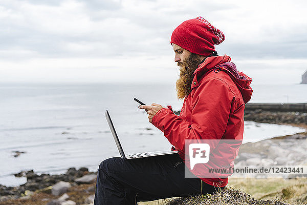 Iceland  North of Iceland  man with laptop sitting near the sea using cell phone