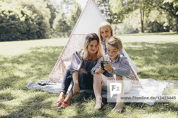 Two young women and a boy looking at cell phone next to teepee in a park