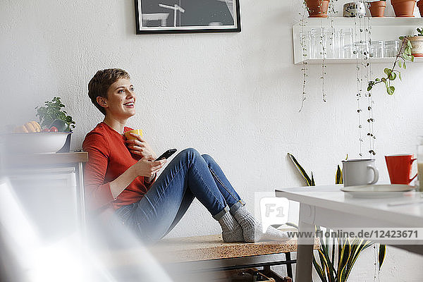 Woman sitting in kitchen  drinking coffee and checking smartphone messages