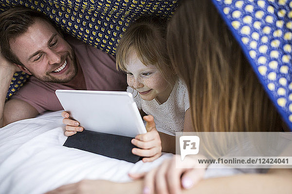 Parents watching something on digital tablet with their daughter under blanket in bed