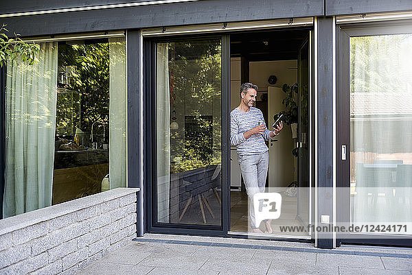 Smiling mature standing at French door at home using a tablet