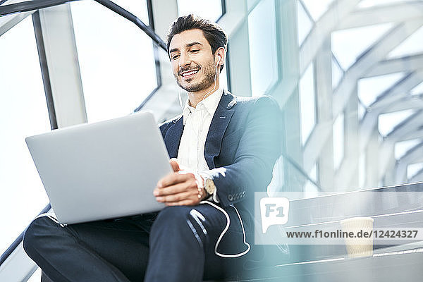 Smiling businessman sitting on stairs wearing earphones and using laptop