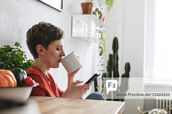 Woman sitting in kitchen  drinking coffee and checking smartphone messages