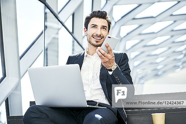Smiling businessman sitting on stairs wearing earphones using cell phone and laptop