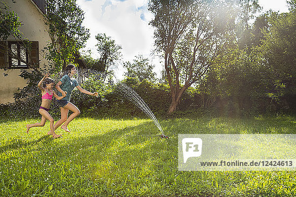 Girl and her little sister having fun with lawn sprinkler in the garden