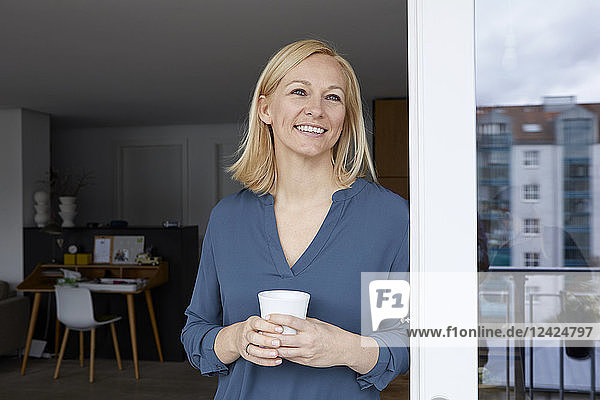 Smiling woman holding cup of coffee looking out of balcony door