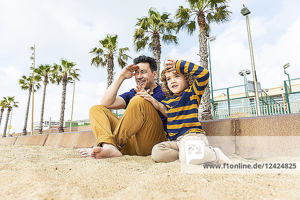 Spain  Barcelona  father and son sitting on the beach looking at distance