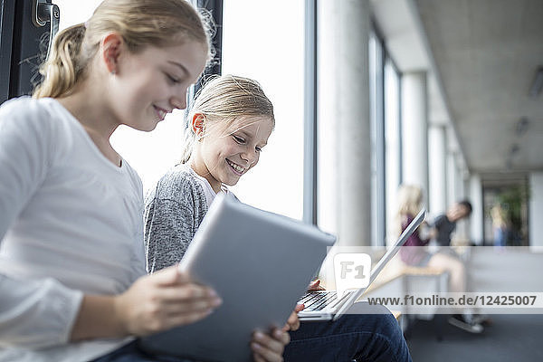 Happy schoolgirls with laptop and tablet on school corridor