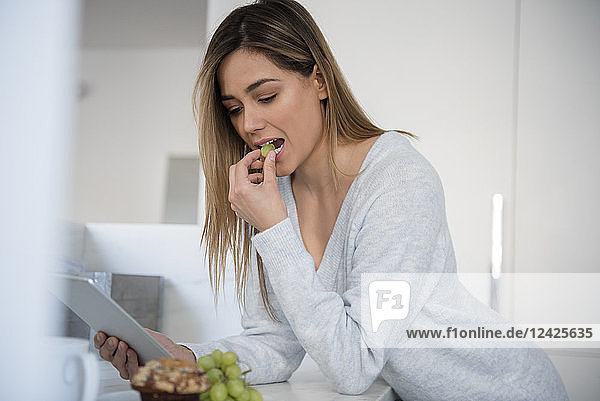 Junge Frau isst weiße Weintrauben und benutzt ein digitales Tablet