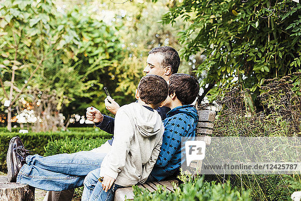 Vater und zwei Söhne (10-11) sitzen auf einer Bank im Park und schauen auf ihr Smartphone