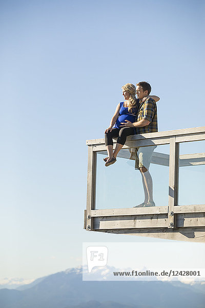 Junges Paar mit schwangerer Frau auf einem Beobachtungsposten gegen den klaren Himmel sitzend