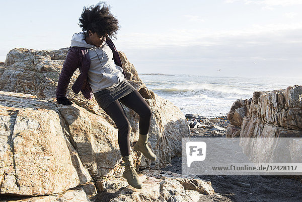 Eine afroamerikanische Frau in einer lila Jacke springt von einem Felsen am Meer herunter