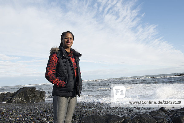 Dreiviertel-Porträt einer lächelnden afroamerikanischen Frau  die auf einem Kiesstrand vor dem Meer steht