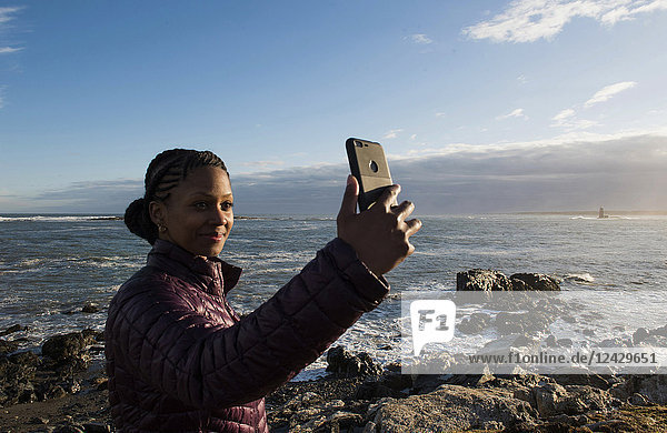Taillenaufnahme einer alleinstehenden afroamerikanischen Frau  die ein Selfie am Meer macht