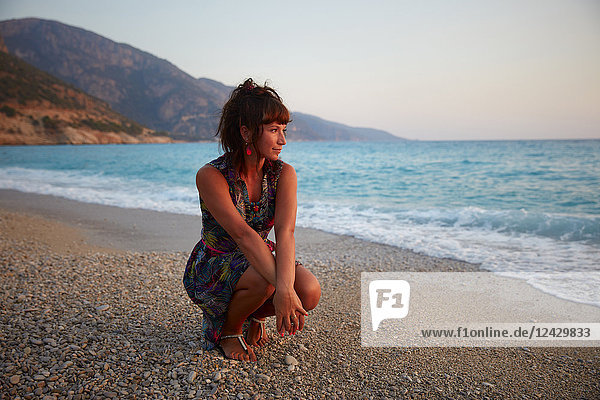 Full length portrait of beautiful young woman crouching on beach at sunset