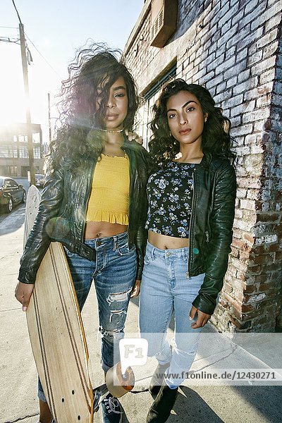 Two young smiling women with long curly black hair standing on pavement  holding skateboard  looking at camera.
